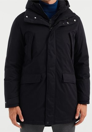 Homme portant un manteau d’hiver noir à capuche avec plusieurs poches zippées et avec rabats, par-dessus un pull à col roulé bleu.