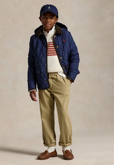 Poika seisoo päällään laivastonsininen tikattu takki, valkoinen villapaita punaisilla raitaviivoilla, beige chinot, ruskeat kävelykengät, valkoiset sukat ja laivastonsininen lippalakki.