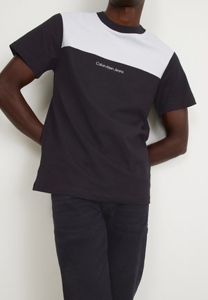 Hombre con camiseta Calvin Klein Jeans en blanco y negro y pantalones negros, de pie con las manos detrás de la espalda contra una pared lisa.