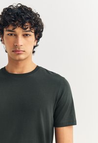 Temno zelena majica z okrolim izrezom in kratkimi rokavi, izdelana iz gladkega blaga, s prilagojeno silhueto in minimalnimi šivi.