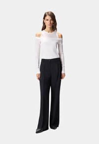 Donna in piedi davanti a uno sfondo uniforme che indossa un top bianco a maniche lunghe con spalle scoperte, pantaloni neri a gamba larga e scarpe nere con punta appuntita.