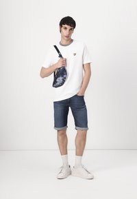 White cotton T-shirt with a yellow logo, dark blue denim shorts, and white sneakers. The individual holds a black and blue tie-dye sling bag.