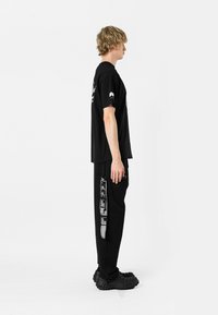 Persona con camiseta negra y pantalones. La camiseta presenta un diseño gráfico; los pantalones tienen franjas laterales con texto impreso y patrones. Calzado de goma.