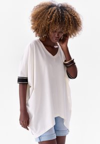 White oversized V-neck top with black striped cuffs, paired with denim shorts. The fabric appears soft and flowing with a relaxed fit.