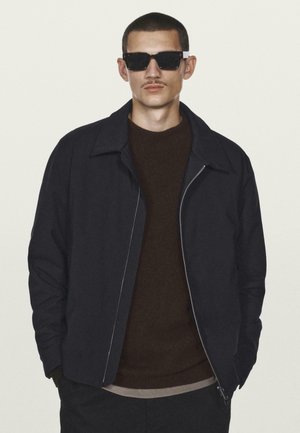 Mand med kort hår og overskæg iført sorte solbriller, mørk jakke over brun sweater, stående mod en lys baggrund.