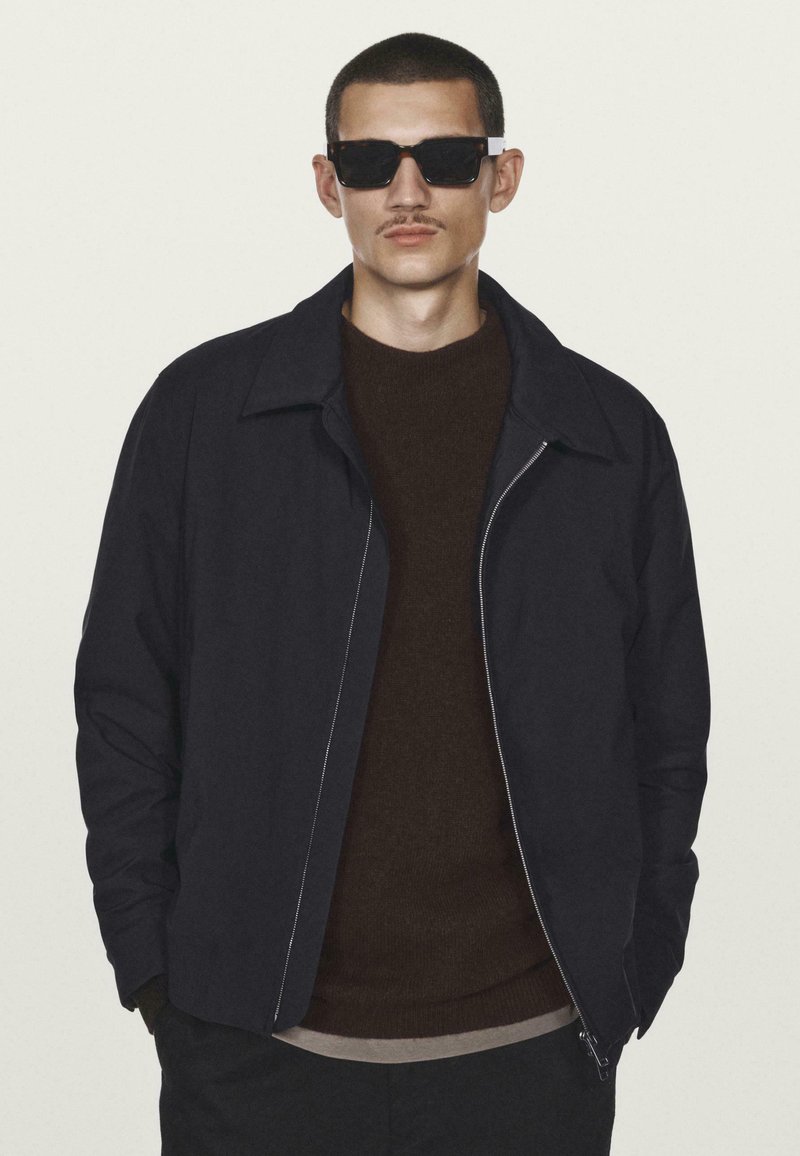 Mand med kort hår og overskæg iført sorte solbriller, mørk jakke over brun sweater, stående mod en lys baggrund.