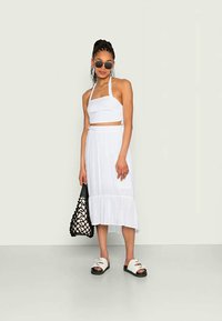 Conjunto branco de duas peças, apresentando um top cortado com alças halter e uma saia fluida, combinado com uma mala com padrão preto e branco e sandálias.