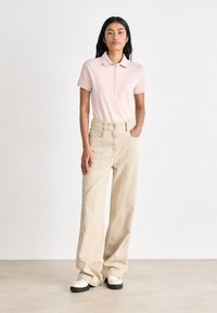 Camisa polo rosa clara de mangas curtas, combinada com calças largas beges de cintura alta e mocassins brancos com solas pretas.