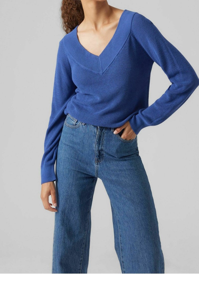 Maglione blu lavorato a maglia con scollo a V profondo e maniche lunghe, abbinato a jeans blu a vita alta e vestibilità ampia. Design semplice e casual.