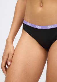 Bañador negro con una cinturilla elástica de color púrpura claro que presenta el logo de "PUMA". La tela parece suave y elástica.
