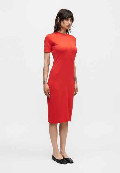 Γυναίκα που φοράει εφαρμοστό κόκκινο φόρεμα μέχρι το γόνατο με κοντά μανίκια, συνοδευόμενο από μαύρα μπαλαρίνια, σε απλό λευκό φόντο.