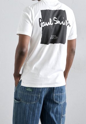 Personne portant un t-shirt blanc avec un carré noir et le texte "Paul Smith" dans le dos, associé à un pantalon en denim bleu rayé avec des poches.