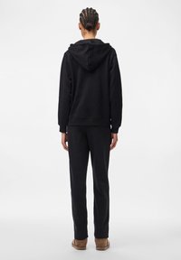 Juodas džemperis su dvigubos medžiagos gobtuvu, kengūros kišene ir laisvu siluetu, derintas su atitinkančiomis juodomis sportinėmis kelnėmis iš minkštos medžiagos.