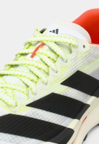 Αθλητικό παπούτσι με ελαφρύ δίχτυ στο πάνω μέρος σε λευκό και διαφανές γκρι, με μαύρες ρίγες, ζωηρά κίτρινα κορδόνια και κόκκινες λεπτομέρειες.