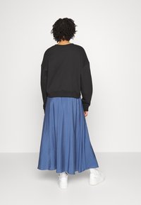 Personne aux cheveux bouclés debout de dos, vêtue d'un haut noir à manches longues, d'une jupe bleue longueur cheville et de baskets blanches sur un fond uni.