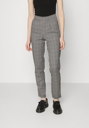 Kvinde iført sorte og hvide ternede bukser med høj talje og forreste folder samt sorte læder loafers, stående mod en ensfarvet baggrund.