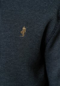 Tamno plava teksturirana tkanina sadrži malu zlatnu izvezenu figuru. Materijal ima karirani uzorak i mekan je na dodir.