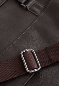 Braune Ledertasche mit strukturierter Oberfläche, ausgestattet mit einem dunkelburgunderfarbenen Riemen und einem Metallverschluss, hervorgehoben durch kontrastierende Nähte.