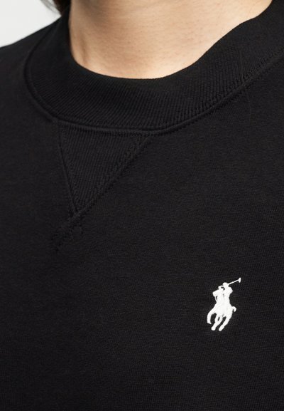 Κοντινή λήψη μαύρου πουλόβερ με στρογγυλή λαιμόκοψη και λευκό κεντητό λογότυπο παίκτη πόλο στο στήθος.