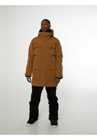 Parka imperméable marron avec capuche, quatre poches avant et boutons-pression. Associé à un pantalon noir et des chaussures noires. Texture lisse.