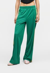 Άτομο που φοράει πράσινο παντελόνι φόρμας φαρδύ με κίτρινες λωρίδες στο πλάι και ροζ και λευκά παπούτσια χωρίς κορδόνια, στέκεται μπροστά σε απλό φόντο.