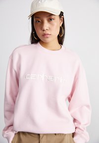 Felpa rosa chiaro con collo a girocollo a coste e logo "carhartt" ricamato. Indossata con pantaloni beige e un cappellino beige. Presenti orecchini a cerchio dorati.