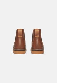 Botas de cuero marrón hasta el tobillo con un acabado suave, paneles laterales elásticos y una suela en contraste color tostado. Presenta un logo de la marca en el talón.