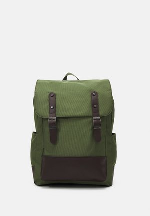 Grüner Canvas-Rucksack mit dunkelbraunen Lederriemen und Bodenteil, Frontklappe und Seitentaschen vor einem einfarbigen hellen Hintergrund.