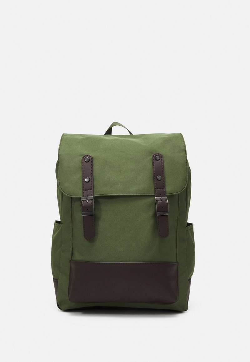 Sac à dos en toile verte avec des sangles en cuir marron foncé, un panneau inférieur, un rabat avant et des poches latérales sur un fond clair uni.