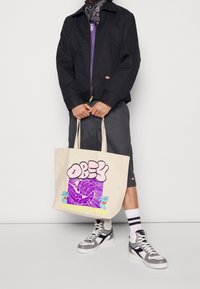 Beige canvas shoppingväska med djärv "OBEY" text i rosa, en lila grafik med tecknade detaljer och slitstarka axelremmar.