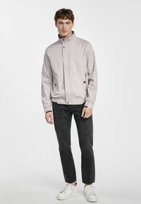 Jasnoszara kurtka z wysokim kołnierzem, zasuwana na zamek, z bocznymi kieszeniami i kontrastującymi brązowymi akcentami, zestawiona z ciemnymi dżinsami i białymi trampkami.