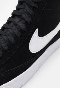 Svart mockasinsneaker med vit Nike-swoosh, med texturerade skosnören och en gummiyttersula. Närbild på designhöjdpunkter och sömmar.