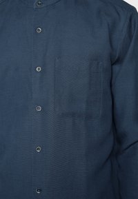 Camisa azul marino con botones, confeccionada en tejido texturizado, que presenta un cuello alto, seis botones y un bolsillo en el pecho.