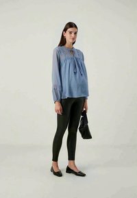 Ljusblå blus med lång ärm och veck samt spetsdetaljer, kombinerad med svarta leggings. Modellen bär en svart handväska och har på sig svarta ballerina.
