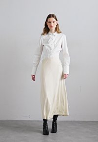 Bílá košile s límečkem, kombinovaná s krémovou dlouhou sukní. Model má na sobě černé, texturované kotníkové boty.