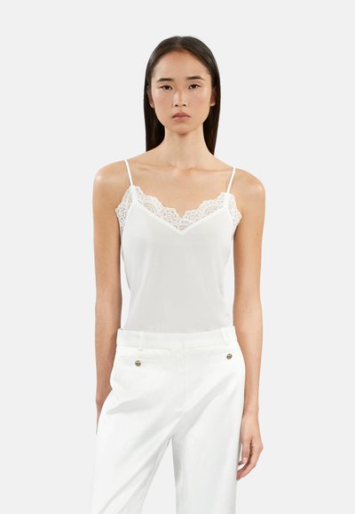 Camisole blanche avec un délicat bord en dentelle au niveau de l'encolure, dotée de fines bretelles réglables. Le tissu a une texture lisse. Associée à un pantalon blanc.