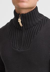 Pull en maille noir avec un col haut à côtes, doté d'une fermeture éclair demi-longue avec un tirette beige et un motif texturé sur les épaules.