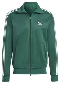 Grön zip-up sportjacka med vit tre-stripes-design på ärmarna, tillverkad av ett slätt material, med en liten vit logotyp och fickor med dragkedja.