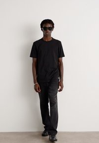 Tricou negru din bumbac, blugi negri skinny și ochelari de soare negri. Modelul pășește pe lângă un perete alb simplu, evidențiind un stil simplu, monocrom.