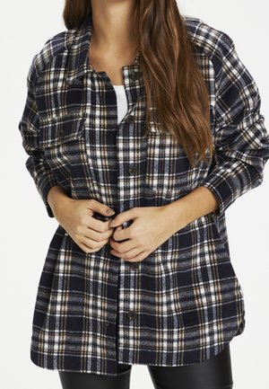 Femme aux longs cheveux boutonnant une chemise à carreaux bleu marine, blanc et beige par-dessus un haut blanc, portant un pantalon noir sur fond blanc.