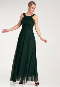 Ciemnozielona, sięgająca podłogi sukienka z dopasowanym gorsetem, szerokimi ramiączkami i zwiewnym tiulowym nałożeniem. Posiada marszczoną tkaninę w talii.
