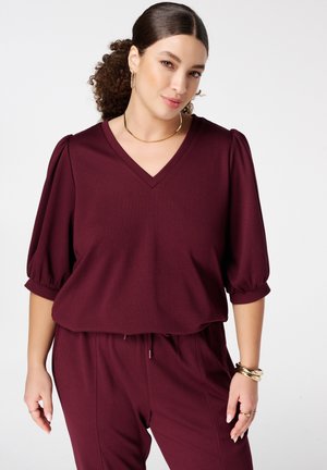 Vrouw met krullend haar in een knot draagt een bijpassende bordeauxrode V-hals top met pofmouwen en losse broek, gecombineerd met gouden sieraden.