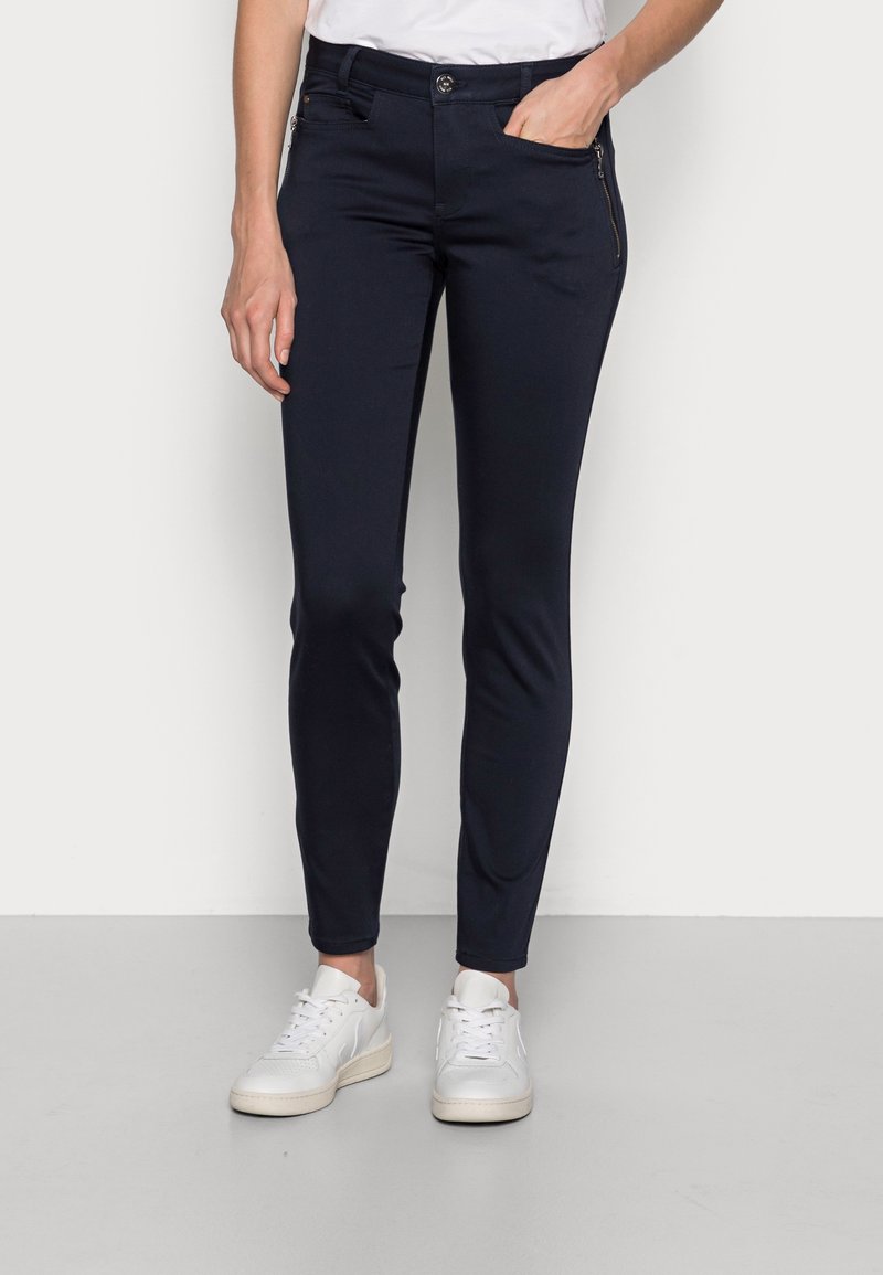 Navy slim-fit kalhoty vyrobené z elastického materiálu, s kapsou na zip, zapínáním na knoflík a drobnými designovými detaily, v kombinaci s bílými teniskami.