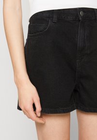 Pantalones cortos de mezclilla negra con un corte clásico, que cuentan con bolsillos frontales y una textura sutil. El dobladillo está bien terminado, complementando el diseño.