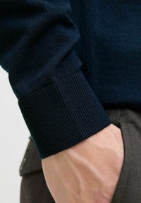 Jūros mėlyna megzta džemperio rankovė su gumuline tekstūra ir lygiu paviršiumi; ranka remiasi į rudą audinį, atskleidžiant odos detales.