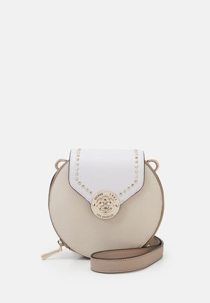 Round beige crossbody bag with white flap, gold stud accents, Guess logo medallion, and adjustable strap on a white background.