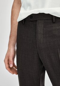 Un primo piano di una mano di un uomo appoggiata vicino a un paio di pantaloni eleganti neri con bottone e passanti per cintura, indossati con una maglietta di colore chiaro.