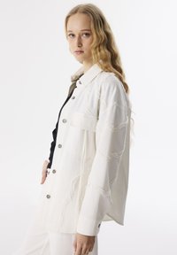 Giacca di jeans bianca con motivi a onde testurizzati, chiusura anteriore con bottoni, tasche laterali e vestibilità rilassata. Abbinata a un top nero sotto.