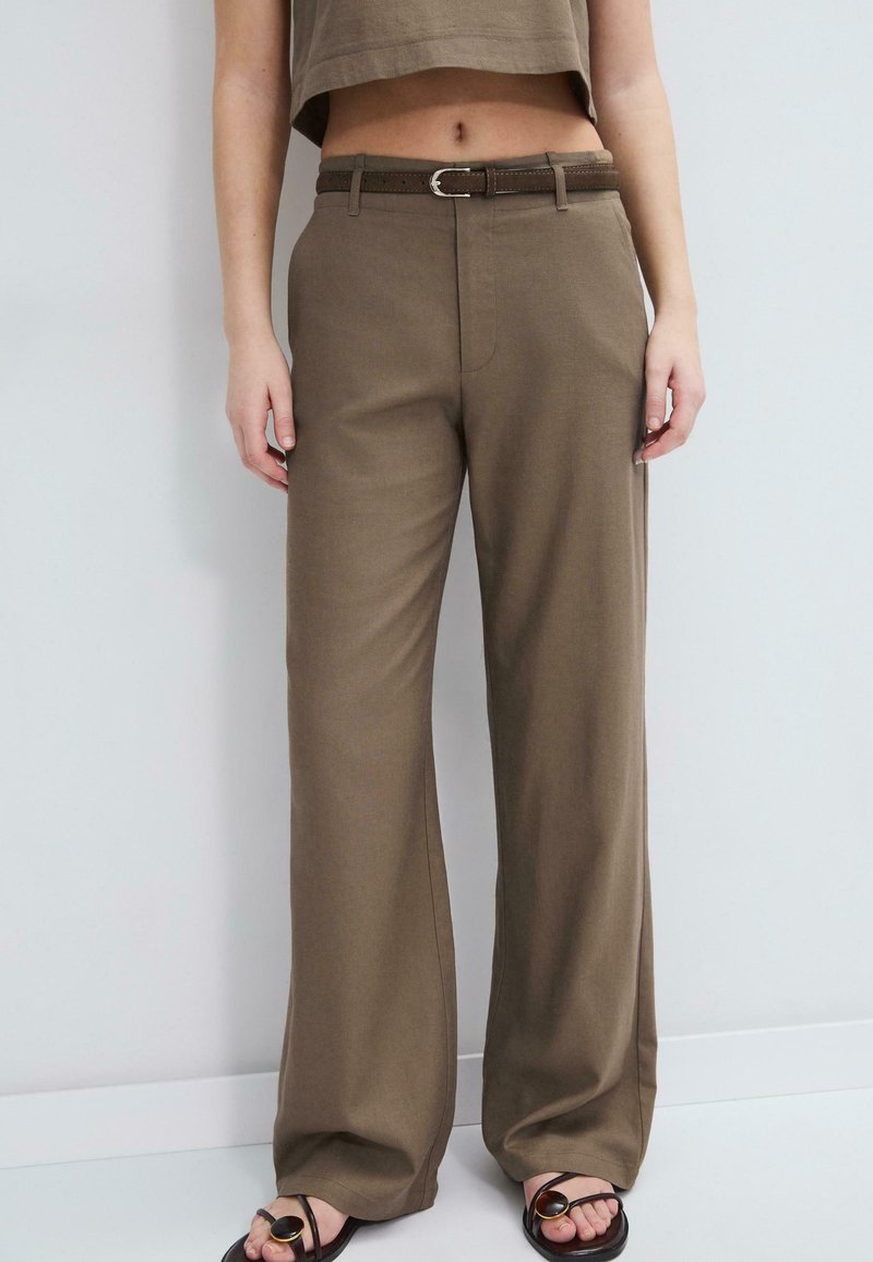 Woman wearing high-waisted, wide-leg brown pants with a matching belt, paired with black sandals, standing against a light wall.