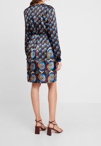 Robe à manches longues, ornée d'un motif sur fond sombre avec des designs bleus et marron. Associée à des sandales à talons bloc avec lanières.